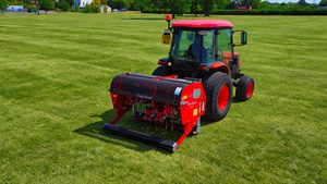 Compact tractor with a red Redexim Verti-Drain deep tine aerator on a freshly mown grass sports field
